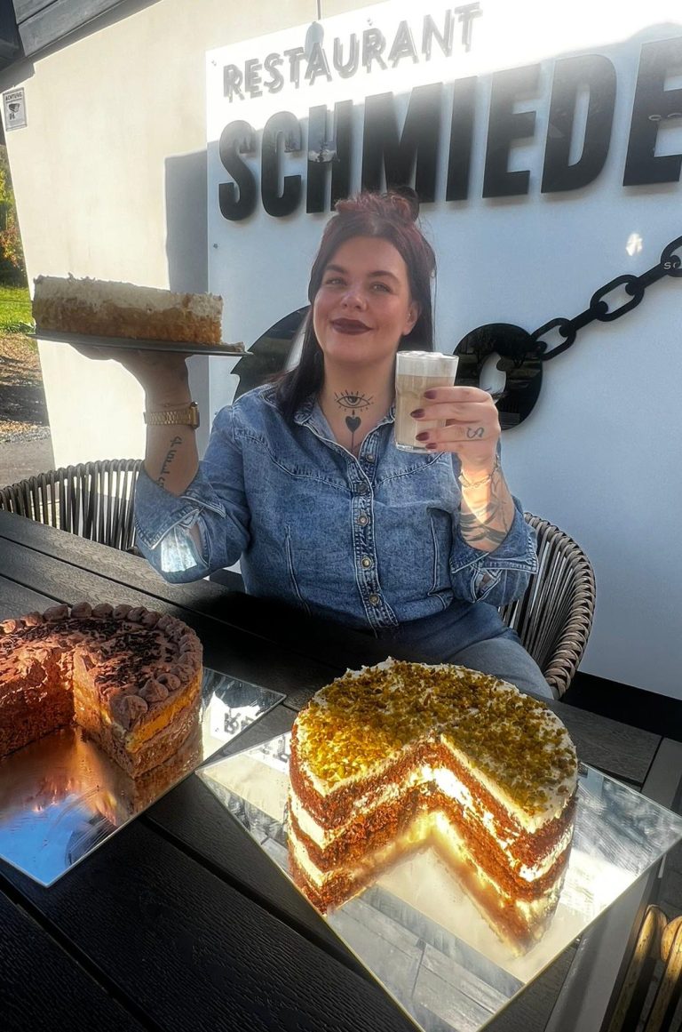 Konditorin Frau sitzt vor einem Schild, hält eine Torte und ein Getränk, mit Kuchen daneben.