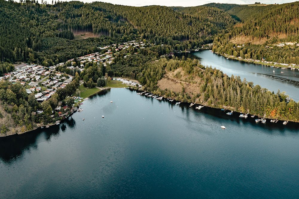 Landschaft mit Fluss, Bäumen und Wohngebieten an den Ufern in einer hügeligen Umgebung.